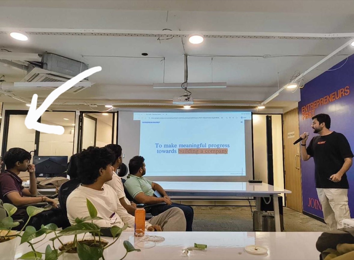 A talk at the Entrepreneurs First office in Bengaluru. A slide reads 'To make meaningful progress towards building a company.' A white arrow points to Divyansh, seated at the far left of the room.