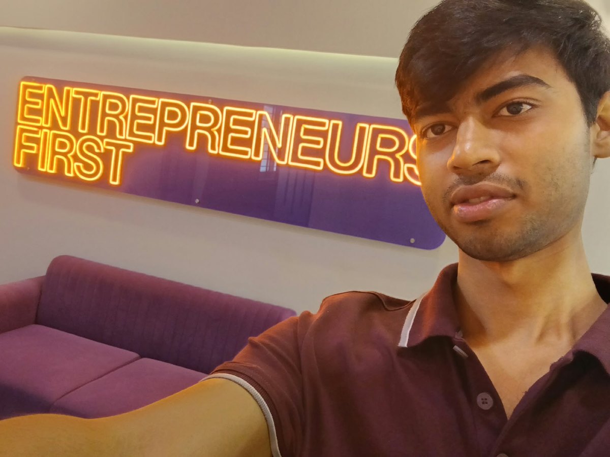 Divyansh Chauhan taking a selfie in front of a glowing orange neon sign reading Entrepreneurs First, mounted on a purple panel above a purple sofa.