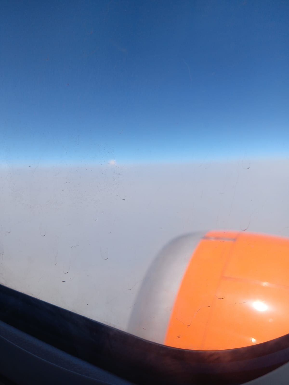 View from an aircraft window at altitude: deep blue sky above a flat white cloud layer, with the orange engine cowling visible below.