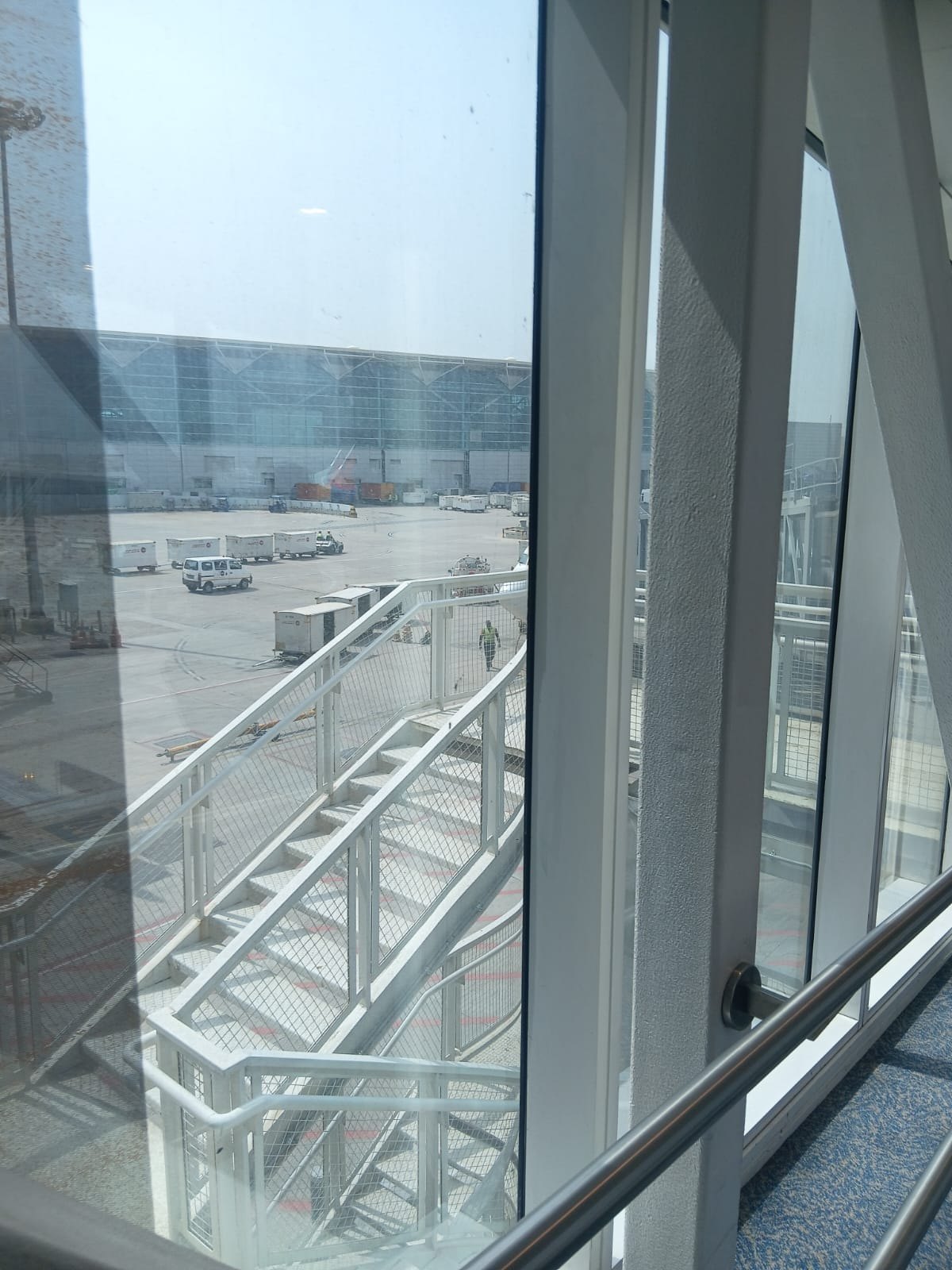 View through an airport terminal window onto the tarmac: jet bridge stairs, service vehicles and baggage containers outside a glass terminal building.